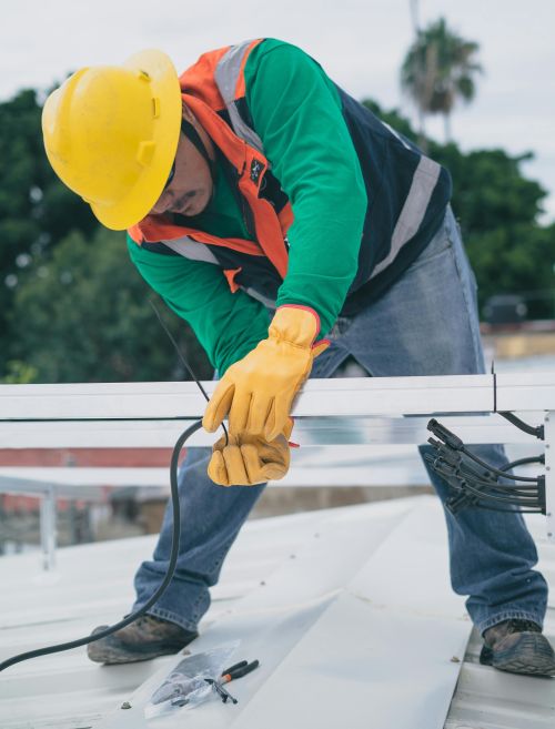 A construction worker wearing PPE installs electrical equipment on a roof.